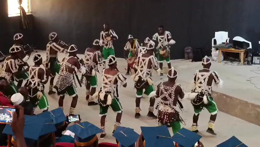 A group of men are performing a dance in front of a seated audience. The dancers are wearing matching traditional outfits including short green shorts, vests with an intricate white and black design, and beaded headdresses. Some of the dancers are playing drums. The audience is seated, wearing graduation gowns and caps, and several people are recording the performance with their phones. The performance is taking place in a large room with black walls and a concrete floor.