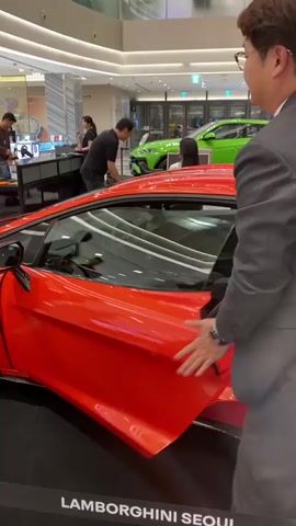 The video captures an indoor luxury car display or event, likely held in a modern, well-lit hall with high ceilings, possibly within a shopping mall or an exhibition space.

The primary subject is a vibrant red Lamborghini sports car, prominently displayed in the foreground. It features sleek lines, black multi-spoke wheels, and distinctive red brake calipers. In the background, another Lamborghini vehicle, a bright green Urus SUV, is also visible. Black barriers with white lettering partially enclose the display area.

Several people are present, mostly adult men, many dressed in dark suits or smart casual attire, suggesting a formal or semi-formal event. Two women, possibly event staff, wearing black uniforms, can be seen in the background near display tables.

The main activity centers around the red Lamborghini. A man in a dark suit walks past the car while looking at his smartphone. He then approaches the front of the red vehicle and proceeds to open its hood, allowing attendees to view the internal components. Other individuals are seen observing the cars with interest, and some are engaged with their phones. The overall atmosphere suggests an exclusive viewing or promotional event for the luxury vehicles.

The visible text on a black barrier at the bottom of the frame clearly reads "LAMBORGHINI SEOUL."
