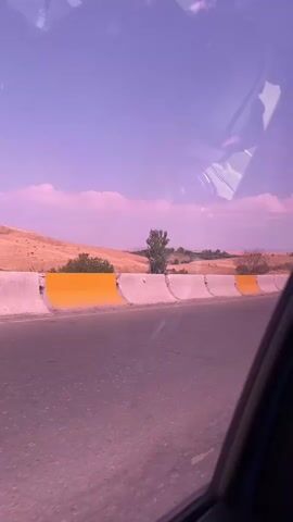 The video shows a view from inside a vehicle looking out at a highway. The sky is a light purple color, and there are scattered white clouds. The landscape on either side of the road is dry and hilly, with sparse vegetation. A concrete barrier with yellow accents runs along the side of the road. Several vehicles, including a white truck with a flatbed and a white car, are seen passing by on the highway. The camera appears to be moving, suggesting the video was filmed from a moving vehicle.