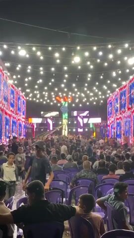 A large crowd of men is gathered outdoors at night for an event. The space is decorated with numerous strings of white lights overhead and vertical LED screens or banners along the sides. The side decorations have a repeating pattern of blue and red lights. In the background, there is a stage with a large screen displaying colorful graphics and a person standing on it. The audience members are seated in purple chairs and appear to be watching the stage. Some people are standing and walking around in the area in front of the stage. The overall atmosphere suggests a concert, festival, or a similar public gathering.