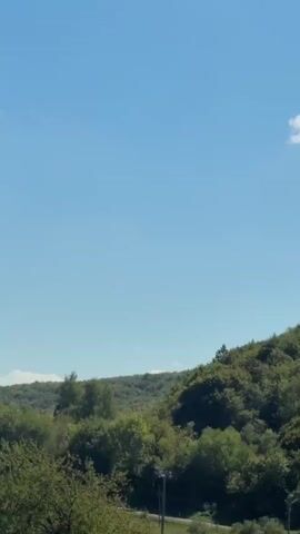 The video is a landscape shot from a moving vehicle, likely a car. The view is of a rural area with rolling green hills covered in trees. In the foreground, there are some bushes and a metal railing. A road is visible in the middle ground, with a white van driving on it. Scattered amongst the trees are a few houses with red roofs. The sky is clear and blue with a few small white clouds in the distance. The overall impression is one of a serene, natural environment.