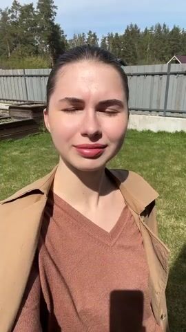 A young woman is standing outside on a sunny day, squinting into the sun. She is wearing a light brown trench coat over a brown V-neck sweater and white sweatpants. Her hair is pulled back in a bun. She turns her head to the side and then looks back at the camera. The background includes green grass, a metal fence, trees, and a house in the distance.