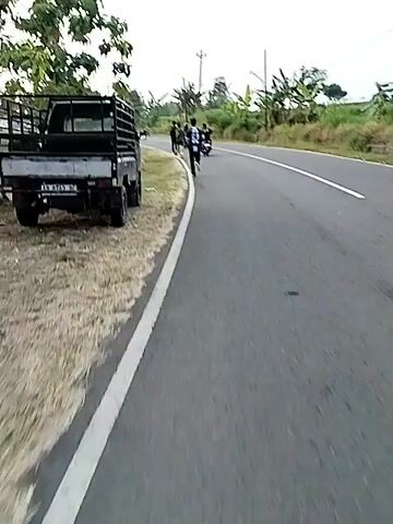 The video shows a man running on the side of a paved road. The man is wearing a dark t-shirt with the number 62 on the back and dark pants. He is running away from the camera, towards a group of people and parked motorcycles on the left side of the road. In the background, there are trees, houses, and power lines. The road has a white line marking the edge. A pickup truck is parked on the left side of the road, near the running man. Another person is running behind the first man, and a motorcycle is passing them on the road. The sky is overcast.