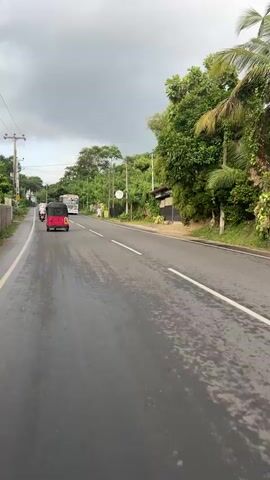 A video begins with a close-up of a wet asphalt road, reflecting a cloudy sky. A red tuk-tuk is seen driving away from the camera on the left side of the road, followed by a white and red bus. The bus passes the tuk-tuk. The road continues with lush green trees and vegetation on the right, and a brown building with black shutters. Power lines are visible overhead. The scene shifts to a close-up of a man wearing a red helmet and sunglasses, looking directly at the camera. He appears to be on a motorcycle, and the road and foliage are visible behind him, slightly out of focus. The road is wet, suggesting recent rain.