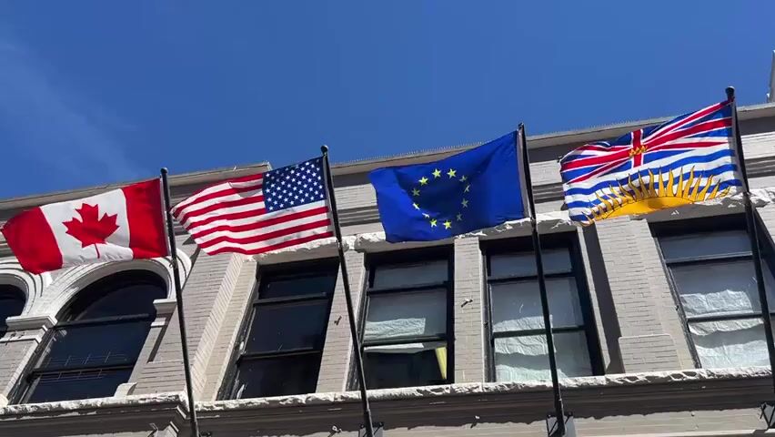 The video shows a close-up, low-angle shot of five flags flying from flagpoles attached to the side of a building. The flags are, from left to right: the Canadian flag, the flag of the United States of America, the flag of the European Union, the flag of British Columbia, and the flag of the city of Victoria. The flags are all in motion due to the wind. The building is light gray and appears to be several stories high with windows and decorative architectural elements. The sky is clear and blue. The video pans slightly to the right, revealing more of the building and the surrounding environment, including some green foliage on the far right. The bottom of the video shows a sign that reads "EXPERIENCE VICTORIA" with several images above it. The lighting suggests it is daytime.