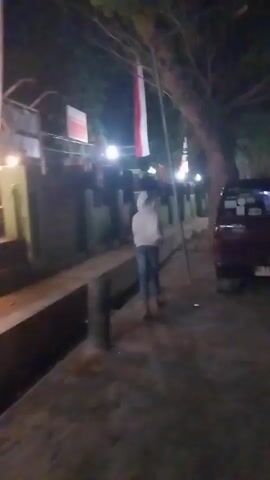 A person wearing a hooded sweatshirt and jeans runs down a dirt path next to a concrete channel. The path is lit by streetlights, and there are red and white flags hanging from poles. A red vehicle is parked on the right side of the path. The video is dark and blurry.