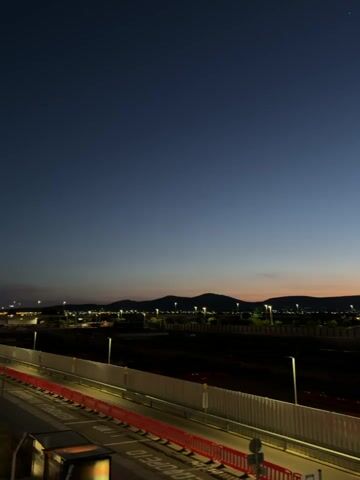 The camera slowly pans across an evening cityscape at dusk. The sky is a gradient of dark blue transitioning to orange near the horizon. In the distance, silhouetted hills and mountains are visible. The city's landscape is punctuated by streetlights and building lights, creating a sparse, twinkling effect. A barrier and a road are visible in the foreground, with construction equipment and materials scattered across the area between the road and the city lights. The view is static for most of the video, with only the camera movement providing any change in the scene.