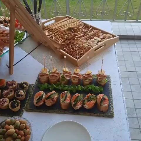 The video shows a table laden with an assortment of appetizers and snacks, likely set up for a buffet or catered event. The table is covered with a white tablecloth, and the background suggests an outdoor setting, possibly a patio or balcony with a railing and some greenery visible.

In the foreground, there are several platters and trays of food. On the left, there's a tray with small tartlets filled with various toppings, including what appears to be seafood, caviar, and some garnished with gold leaf. Next to this, a small white plate holds dried fruit or preserved vegetables, possibly tomatoes and figs, and a few nuts. To the right of the tartlets, there's a slate platter with more small bites, including bread slices topped with salmon, cream cheese, and herbs, as well as what looks like miniature wraps or rolls.

Further back, a large glass platter showcases a charcuterie and cheese board, generously arranged with a variety of sliced meats like salami and prosciutto, cheeses, and garnishes of lettuce and herbs. Behind this, a wooden tray is filled with an assortment of breadsticks, crackers, and small savory pastries or pretzels. To the left of the charcuterie board, there's another platter with more appetizers, some skewered with cherry tomatoes and olives.

The lighting suggests it's daytime. There are no people clearly visible in the frame, though a hand might be seen briefly in the very first frame on the far left, reaching towards the food. The overall impression is one of abundance and variety, typical of a party or event where finger foods are served.