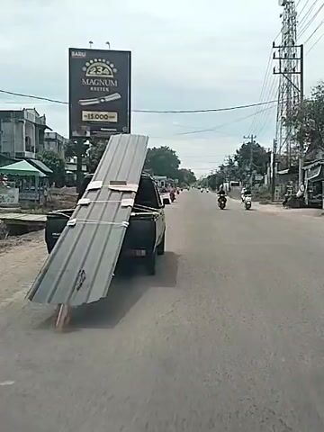 A black pickup truck drives down a gray asphalt road during daylight. The truck bed carries a stack of gray corrugated metal sheets secured with straps and wooden planks, extending far beyond the vehicle's rear. A piece of cardboard is wedged under the overhanging sheets. Other motorbikes and vehicles are visible on the road ahead. The roadside features buildings, trees, and power lines. A large advertisement for "Magnum Kretek" is visible on a billboard to the left of the road. The sky is overcast. The camera follows the truck from behind.