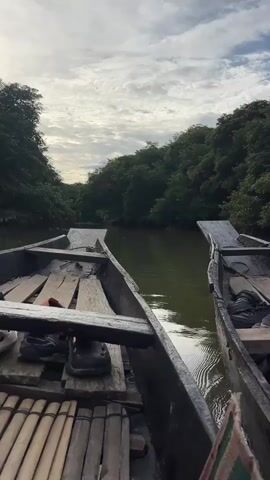 The video shows a view from the front of a wooden boat moving down a narrow river surrounded by lush green trees. Two similar wooden boats are visible, one to the left and one to the right of the camera's boat. The boat on the left has a person's legs and feet visible, wearing black shoes and a piece of orange fabric. The boat on the right also has shoes visible. The sky above is cloudy with patches of blue peeking through. The water is dark and reflects the sky and trees. The overall scene depicts a serene boat ride through a natural waterway.