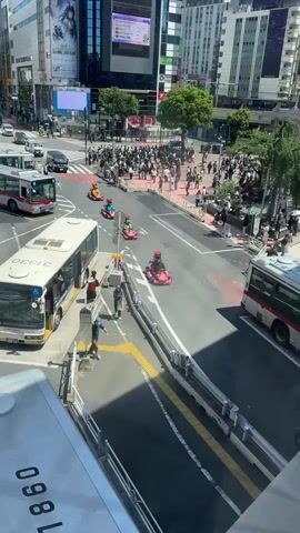 The video shows a busy street in Japan with several buses, cars, and pedestrians. In the middle of the street are four people driving small, red go-karts and dressed as characters from the video game Mario Kart. They are driving in a line, moving from right to left in the frame. The street is lined with sidewalks and tall buildings. Many people are gathered in the crosswalks. The video is taken from an elevated position, possibly from a building or bridge.