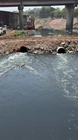 The video depicts a heavily polluted river with dark-colored water flowing through two large pipes into the main body of water. Construction is taking place on the banks of the river. A road or bridge is visible above the water, supported by large concrete pillars. An excavator and other construction equipment can be seen in the background. There is garbage present near the construction area.  The water in the river is visibly turbulent where it exits the pipes. The overall scene appears to be one of environmental degradation due to pollution.