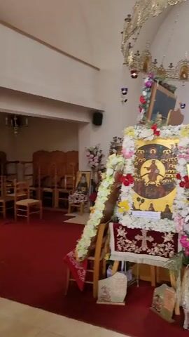 The video shows the interior of a church. In the foreground, a large, ornate icon is displayed on an easel and decorated with flowers. The icon depicts a religious scene. To the left, another icon is visible, and to the right, there are wooden pews and part of a wooden iconostasis. A large, multi-tiered chandelier hangs from the ceiling, adorned with many candles and religious images. The walls are white, and the floor is covered in red carpet. The lighting is dim, with sunlight filtering in from windows at the top. The camera pans slowly across the scene.