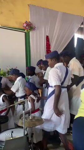 A group of young people in a church perform music, likely religious, with an energetic and coordinated style. They are dressed in white and beige uniforms with blue accents, and wear blue caps. The scene includes a drum set, which one young person plays, and some of the others dance and sing. A banner with religious text is behind them, with a projection screen to the side. The scene has decorations including red tinsel, and the room has a simple, functional, decorated interior.