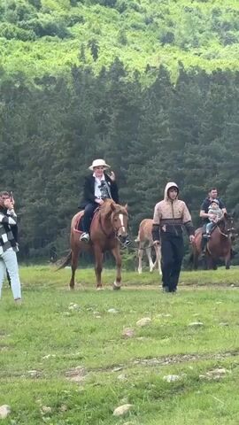 The video shows a grassy field with a backdrop of dense green trees. Four adults and one child are visible. One adult is riding a brown horse and wearing a white hat, a black blazer, and jeans. Another adult is leading a light brown foal. A third adult is riding a brown horse and holding a small child in front of them. A fourth adult is walking on the left side of the frame. The person on the horse wearing the white hat appears to be gesturing with their hand. The horses and people are relatively close together and moving.