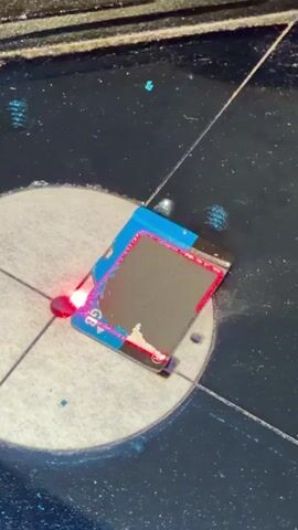 A close-up shot shows a rectangular object, likely a circuit board or similar electronic component, secured on a metallic surface marked with concentric circles and radial lines. A bright red laser beam is actively engaged with the object, creating a visible point of light and emitting faint trails of red light as it moves across the surface. The object appears to have blue tape attached to one side and a metallic gray surface that is being affected by the laser, showing signs of ablation or etching. The laser appears to be controlled by an external mechanism, indicated by its focused beam and controlled movement.
