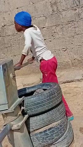 A young woman with a blue head covering operates a hand-powered water pump next to a brick building on a dusty, sandy patch of ground. The pump has a silver metal housing and a metal spout. The video pans to a wider shot of a dusty area lined with trees and low brick walls. Several women in colorful clothing are bent over large tubs, apparently washing clothes. Other people walk along the path in the background. Dogs are also visible roaming the area.