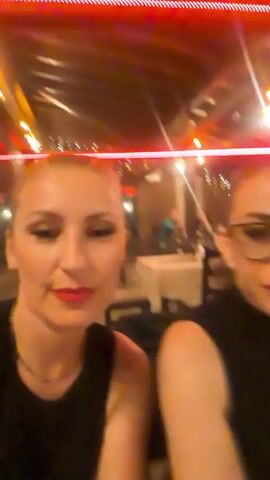 The video captures two women posing for a selfie in what appears to be a restaurant or event space with ambient lighting. The woman on the left, with blonde hair pulled back, wears a black sleeveless top and a delicate necklace. She is smiling warmly. The woman on the right has dark hair tied in a ponytail, wears glasses, and a black turtleneck top. She also smiles, with bright red lipstick visible on both women. The background shows other patrons seated at tables, suggesting a social gathering. String lights are visible overhead, contributing to the warm, festive atmosphere.
