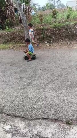 Two children are playing outside on a paved surface. The child on the left is sitting on a toy vehicle that resembles a tank. The child on the right is walking away from the camera. The background consists of trees, vegetation, and a pile of debris. The lighting suggests it is daytime.