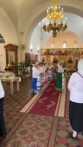 A video shows the interior of a church. In the foreground, the floor is tiled in a diamond pattern with a red rug runner adorned with a scrolling design. To the left, a table is set with food items. In the middle ground, a group of women wearing traditional white dresses with colorful embroidered patterns and floral wreaths on their heads stand in two lines, facing the altar. To the right, another woman in a white top and black skirt stands with her back to the camera. The background features an iconostasis with numerous religious icons. A large, ornate golden chandelier hangs from the arched ceiling. The overall atmosphere appears to be solemn and ceremonial.
