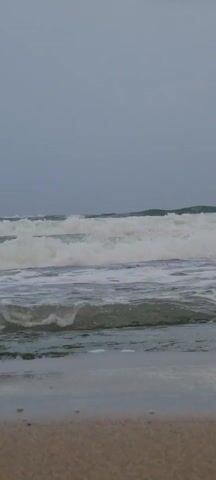 The scene shows a beach on an overcast day. The waves are choppy and covered in white foam. The sky is a uniform grey. There is no visible human presence. The sand is a light tan color.