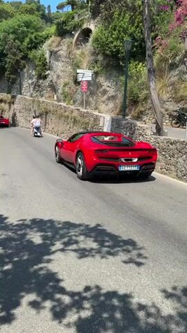 The video shows a red Ferrari driving up a road. The road is narrow and curves to the left. There are trees and bushes on the side of the road, as well as a stone wall. A scooter is also visible on the road, ahead of the Ferrari. There are two people on the scooter. Two people are walking on the sidewalk to the left of the road. The sky is blue and there are some clouds. The sun is shining.