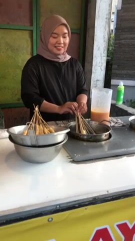 A young woman wearing a light brown hijab and a black long-sleeved shirt is standing behind a food stall. She is smiling and appears to be preparing food. In front of her are two metal bowls filled with skewers of what appear to be fried food items. She is holding a bundle of skewers with both hands, dipping them into a pot of sauce. The stall has a white countertop and a yellow banner with red lettering along the bottom. Behind her, there is a large clear plastic jug filled with orange sauce and a green bottle. The scene is set outdoors, with a wall made of colorful tiles and stone in the background. The lighting suggests it is daytime, possibly late afternoon or early evening. The woman's expression conveys a friendly and welcoming demeanor. The food stall appears to be a street food vendor.
