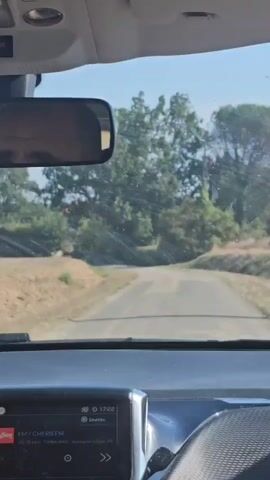 The video shows a view from inside a car driving on a paved road through a rural, tree-lined area. The driver's reflection is visible in the rearview mirror. The car's dashboard display shows the time as 17:22 and indicates the radio station FM CHERIEF M. The sun is shining, casting light and shadows. The road appears to be a single lane in each direction. Trees and vegetation line both sides of the road.
