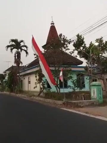 A low-angle, slightly shaky video captures the side of a mosque with a distinct tiered, conical roof topped by a small dome. Two Indonesian flags, red and white, are prominently displayed. The larger flag hangs diagonally across the left side of the frame, obscuring part of the mosque. A smaller flag is mounted on a pole near the right side of the mosque. The mosque itself has white walls with light blue trim and a colorful striped band just below the roofline. Several windows are visible, including small green-barred vents and larger windows with dark frames. A palm tree stands to the left of the mosque, and other trees and vegetation are scattered around the building. The ground in front of the mosque is dirt and grass, with a paved road in the foreground. The sky is overcast. A sign is visible on the left wall of the mosque, but the text is unreadable. The video appears to be filmed from a moving vehicle. There are no people visible in the video.