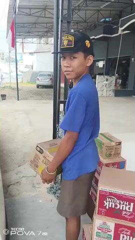 A young boy wearing a blue t-shirt and brown shorts, a black baseball cap, and flip-flops is holding a cardboard box. He is standing in front of a stack of boxes, some of which are labeled "Pucuk Harum". To his right, a woman in a white headscarf and black jacket is looking at the camera. Behind them, there is a building with a blue garage door and motorcycles parked in front. An Indonesian flag is visible on a pole to the left of the frame. The ground is paved and appears to be an outdoor area, possibly a storefront or warehouse. The boy is interacting with someone or something off-camera to his left.