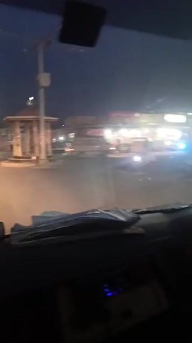 The video is taken from the inside of a vehicle, likely a car or truck, driving on a road at night or dusk. The camera is positioned on the dashboard, providing a driver's point of view. The vehicle approaches a roundabout with a structure in the center that appears to be a pavilion or small monument. A person is seen standing near the roundabout. The surroundings consist of buildings and shops along the roadside, illuminated by artificial lights. As the vehicle passes the roundabout, it continues down a road lined with streetlights and trees on either side. Other vehicles, including a bus, are visible ahead. The overall impression is of an urban or semi-urban environment at twilight or night.