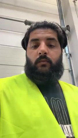 A man with dark hair and a beard is wearing a bright yellow safety vest over a black t-shirt. He is standing in front of a large, off-white industrial door that appears to be a loading bay or warehouse entrance. Above the door, a large spring mechanism is visible. The man is looking directly at the camera and speaking. His mouth is open in surprise or shock. A small white sign with the number "11" is visible on the door behind him. A red logo with white text "log.in" and "expres" is on the right side of his vest. The setting appears to be a warehouse or industrial facility. The lighting suggests it is daytime.