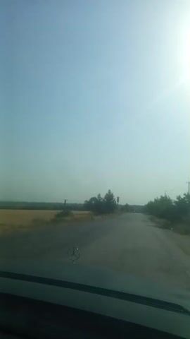 A car drives on a road through a rural area. The sky is clear and blue, and the sun is shining brightly. There are trees and fields on either side of the road. The road is paved and appears to be in good condition. A Mercedes-Benz logo is visible on the hood of the car. The video is taken from the perspective of the driver.