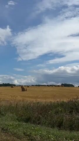 The video captures a wide shot of a rural landscape. A field of harvested wheat is dotted with numerous round hay bales. The field is mostly golden brown, indicating it's been recently cut. In the foreground, there is a strip of green grass and vegetation. The sky above is a bright blue with scattered white and gray clouds. The scene appears to be a sunny day. There are no people or animals visible in the frame. The camera is stationary, and the video is brief, showing minimal change in the scene over its duration.