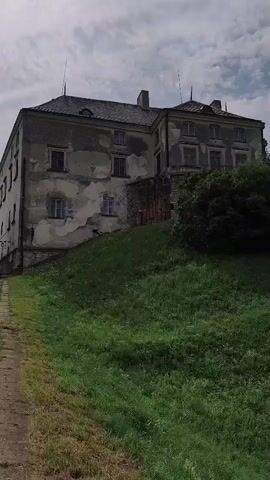 The video pans across a green grassy hill with a large, weathered stone building at the top. The building appears to be an old castle or manor, with crumbling plaster and multiple windows. Trees and bushes are visible around the building and on the slope of the hill. The sky above is cloudy. The camera moves from left to right, showcasing the building and the surrounding landscape.