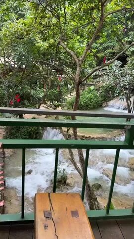 The video shows a view of a waterfall and lush greenery from a balcony. A wooden bench is visible in the foreground, and a railing of a balcony with green bars runs across the bottom of the frame. The waterfall cascades over rocks into a turquoise pool. Trees and dense foliage surround the area, with some red decorations hanging from branches. Stairs are visible in the background, suggesting a path or access to the waterfall area. The water appears clear and the environment is natural and serene.