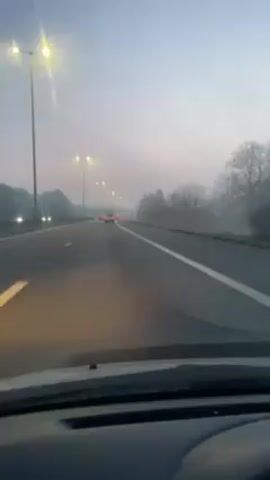 The video shows the view from inside a car driving on a highway at what appears to be dawn or dusk. The sky is a pale gray, and there is fog or mist visible, particularly in the distance. Streetlights illuminate the road, which has multiple lanes in each direction separated by a median.  Several cars are visible on the road ahead, their taillights glowing. The landscape on either side of the highway is mostly trees and vegetation, partially obscured by the fog. The camera is stationary within the car, focusing on the road ahead.
