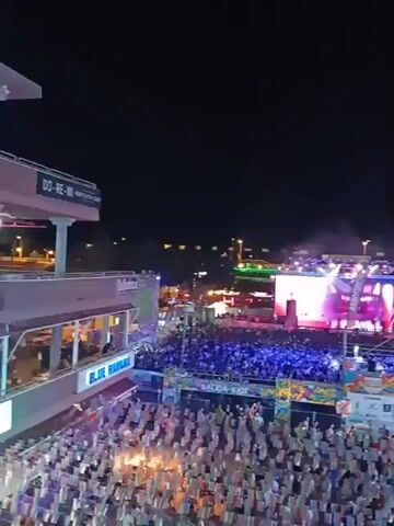 The video shows a large outdoor concert at night. A large crowd of people is packed together in front of a stage with bright lights and video screens. There is a building with a sign that says "Blue Bar & Lounge" to the left of the frame. In the distance, there are buildings and streetlights. The music is loud and energetic, and the crowd is cheering and dancing. There are also flags and banners with different logos, including "ABSOLUT" and "Freedom". The camera is positioned high above the crowd, providing a wide view of the entire event.