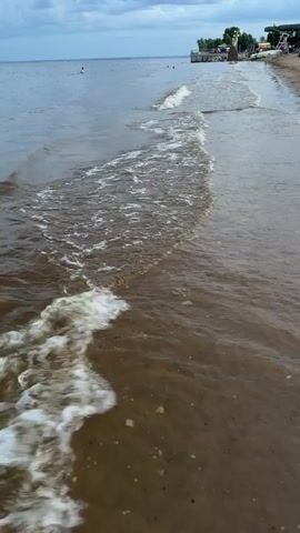 The video shows a beach with waves gently rolling in. The water is a murky brown, and there is some foam on the surface. In the distance, there are a few people swimming in the water. The sky is cloudy, and the overall atmosphere is calm and peaceful. There is a small structure or pier visible on the right side of the frame, with some people standing on it. The beach itself is sandy, with some scattered debris.
