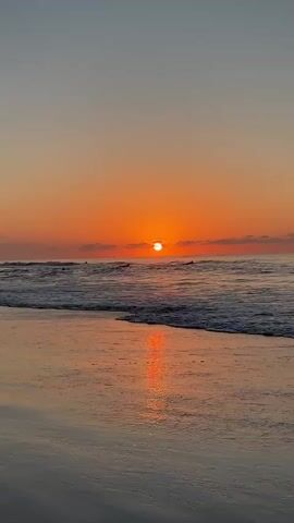 A vertical video captures a tranquil beach scene at sunset. The sun, a bright orange orb, is setting over the ocean, casting a warm glow on the water and the sky. Gentle waves lap at the sandy shore, reflecting the vibrant colors of the sunset. Several silhouetted figures can be seen in the water, possibly surfers, enjoying the last moments of daylight. The sky transitions from a soft orange near the horizon to a pale blue overhead, with a few wispy clouds adding texture to the scene. The overall atmosphere is peaceful and serene.