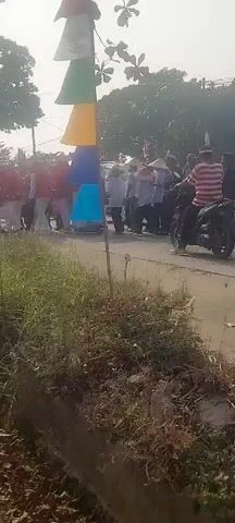 The video shows a street scene with several people on motorcycles. The motorcycles are moving forward in a line on a paved road. The people on the motorcycles are wearing casual clothing. Some of the people are wearing hats. There are trees and buildings in the background, and some flags are visible. The overall scene appears to be a procession or parade. The lighting suggests it is daytime.