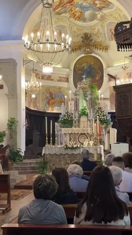 A high-angle interior shot depicts a group of people attending a religious service in a church. Rows of people, seen from the back, sit on dark wooden pews, facing the altar. The altar is elaborately decorated with flowers, candles, and a statue of the Virgin Mary, illuminated by a green light.

Above the altar, there is a large oval painting of a religious figure, and to the sides are smaller murals on the walls. The church is well-lit by several chandeliers, each adorned with numerous light bulbs. A wooden pulpit stands to the right of the altar. The architecture is classical, with an arched ceiling decorated with frescos. The scene is still and quiet, capturing a moment of reverence during the service.