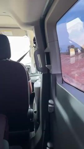 The video, captured from the back seat of a moving vehicle, shows the back of a person's head and shoulders. The person is wearing a dark shirt and a seatbelt. The vehicle is moving on a paved road, and a white van is visible ahead. In the distance, a highway overpass is visible, and the sky is bright and clear. The vehicle's interior is light-colored, with a handle visible on the right side.