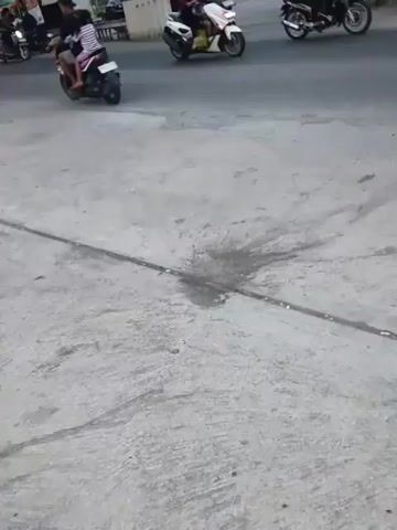 A yellow truck is driving down a paved road. There are several motorcycles on the road as well, some with riders and some without. The area appears to be outdoors and possibly a public road. The ground in the foreground is cracked concrete with a metal strip running across it. There are some dark stains on the concrete near the metal strip. In the background, there are buildings, trees, and power lines. The sky is overcast.