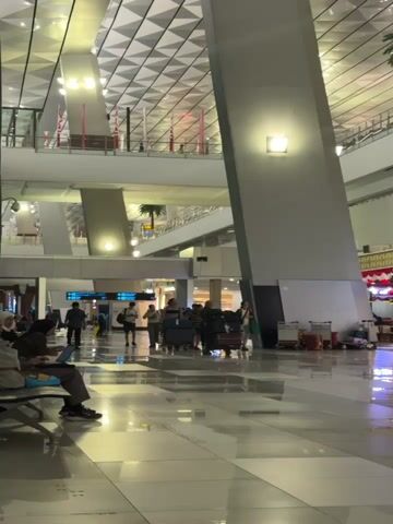 The video shows a wide shot of an airport terminal. A person is sitting on a bench in the foreground on the left, looking at a laptop. In the middle ground, several people are walking with luggage carts. The terminal features a high, geometric ceiling with lighting, large support pillars, and a palm tree is visible in the background. Several food stalls are also visible in the background. The floor is polished and reflects the lights and architecture.