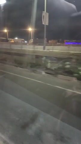 At night, a blue car with white sidewall tires is seen moving to the right along a roadway. The video is shot from a moving vehicle. Streetlights illuminate the scene, and a raised highway section is visible in the background, with other vehicles parked there.