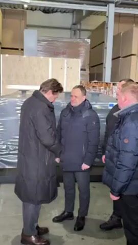 The video captures a scene in what appears to be a warehouse or storage facility. Four men are present, three are visible, and the fourth man is partially visible, they are standing and appear to be conversing. Two of the men are shaking hands while making eye contact. The man on the left is wearing a long black coat and brown shoes. The second man is wearing a navy puffer jacket, dark pants, and black shoes. The third man is also wearing a puffer jacket. Behind the men are large, shrink-wrapped items. Some crates are visible in the background. The location is well lit.
