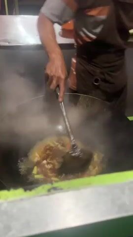 The video shows a close-up shot of a person cooking in a large wok. The scene appears to be an outdoor food stall or a kitchen with a stainless steel counter in the background. The lighting suggests it is nighttime or a dimly lit indoor space. The person, wearing an apron, is actively stirring the contents of the wok with a large ladle, which is generating a significant amount of steam. Ingredients are visible in the wok, appearing to be a mixture of noodles, vegetables, and possibly meat, all coated in a yellowish sauce. In one segment, the person is seen pouring a dark liquid from a bottle into the wok, likely a sauce or seasoning. The overall atmosphere is that of a bustling food preparation environment, emphasizing the dynamic process of stir-frying. There is a package with Indonesian text on it in the background, "Indomie Goreng," which is a popular instant noodle brand in Indonesia. The setting is implied to be Bebekan, Indonesia, based on the provided context.