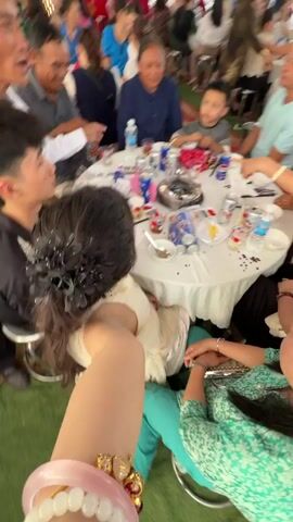 This video captures a lively social gathering, likely a celebratory event, taking place in Bình An, Vietnam. The scene is set outdoors or under a large open-sided tent, with a green ground cover (possibly artificial turf) and sections of red carpet. It appears to be daytime, with ample natural light.

The camera is held from a first-person perspective, belonging to someone seated at a round table. The arm of the cameraperson, adorned with a pink bracelet, a gold and jeweled bracelet, and a watch, is visible in the foreground. The individual is wearing a light-colored (possibly white or cream) dress, and their long dark hair, decorated with black floral pins, is also visible.

Around the table, several individuals are seated, including men, women, and at least one child. They are engaged in eating, drinking, and socializing. On the white-cloth-covered table, there are various dishes, small cups, bowls, spoons, and numerous cans of what appear to be soda (some resembling Pepsi cans). A hotpot or heating element is visible in the center of the table, suggesting a shared meal.

Notable individuals at the table include:
*   A woman in a green patterned top and turquoise pants, seated with her back to the camera.
*   A woman in a white top, actively reaching across the table for a drink can.
*   An older man in a white shirt and a younger man in a black shirt, both looking at other people.
*   An older woman in a blue top.
*   A young boy in a grey shirt, eating from a bowl with a spoon.

In the background, many more people are visible at other tables, some standing, some seated, indicating a large attendance. The atmosphere seems festive and communal, with an upbeat song playing as background music. The event appears to be a joyful occasion, possibly a wedding reception, family reunion, or community feast.
