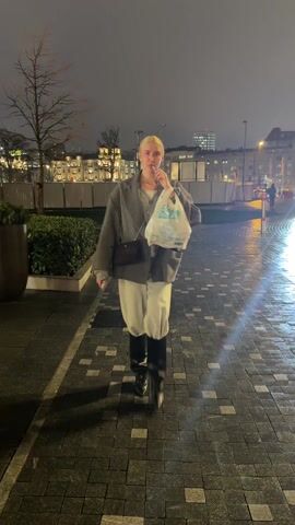 A woman with blonde hair pulled back into a ponytail walks towards the camera on a wet, brick-paved sidewalk at night. She wears a grey oversized blazer, a light-grey sweater, light-colored pants tucked into black boots, and a small brown purse slung across her shoulder. She holds a white plastic bag in one hand and a slim, vape-like device in the other, periodically bringing it to her mouth. The background features manicured hedges, bare trees, and buildings illuminated by streetlights and internal lighting.  A shop front with glass doors and colorful artwork inside is visible briefly to her right. The overall lighting is somewhat bright due to the streetlights.